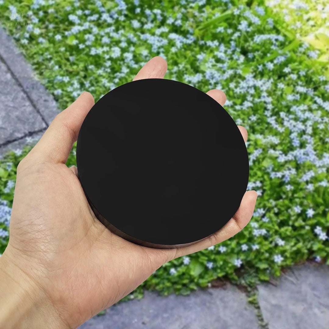 A person holding a 3.94-inch scrying mirror with an obsidian surface. The background shows a flower-filled green area and a stone path